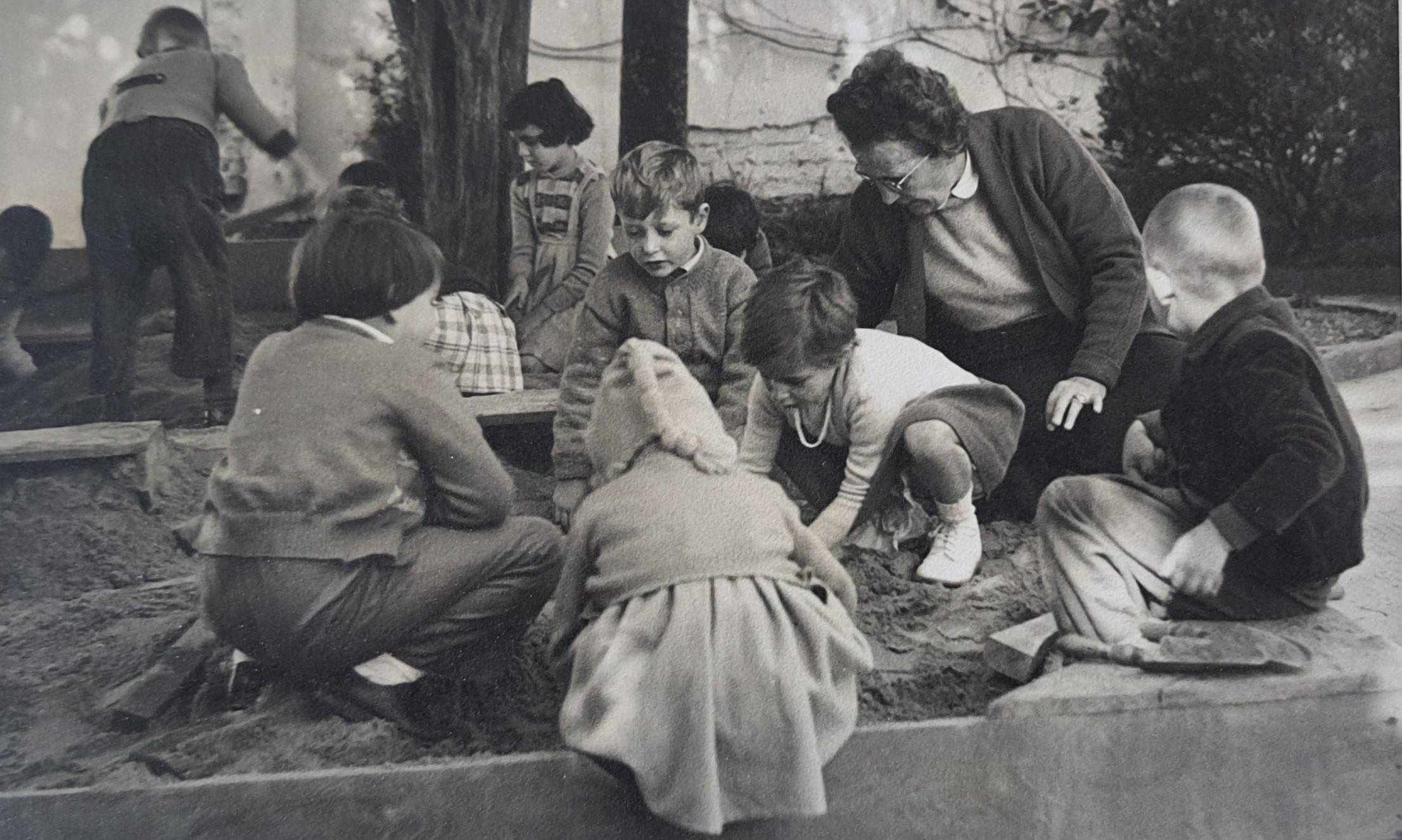 Sra. Ida Ulrich e crianças do jardim de infância brincando na caixa de areia.