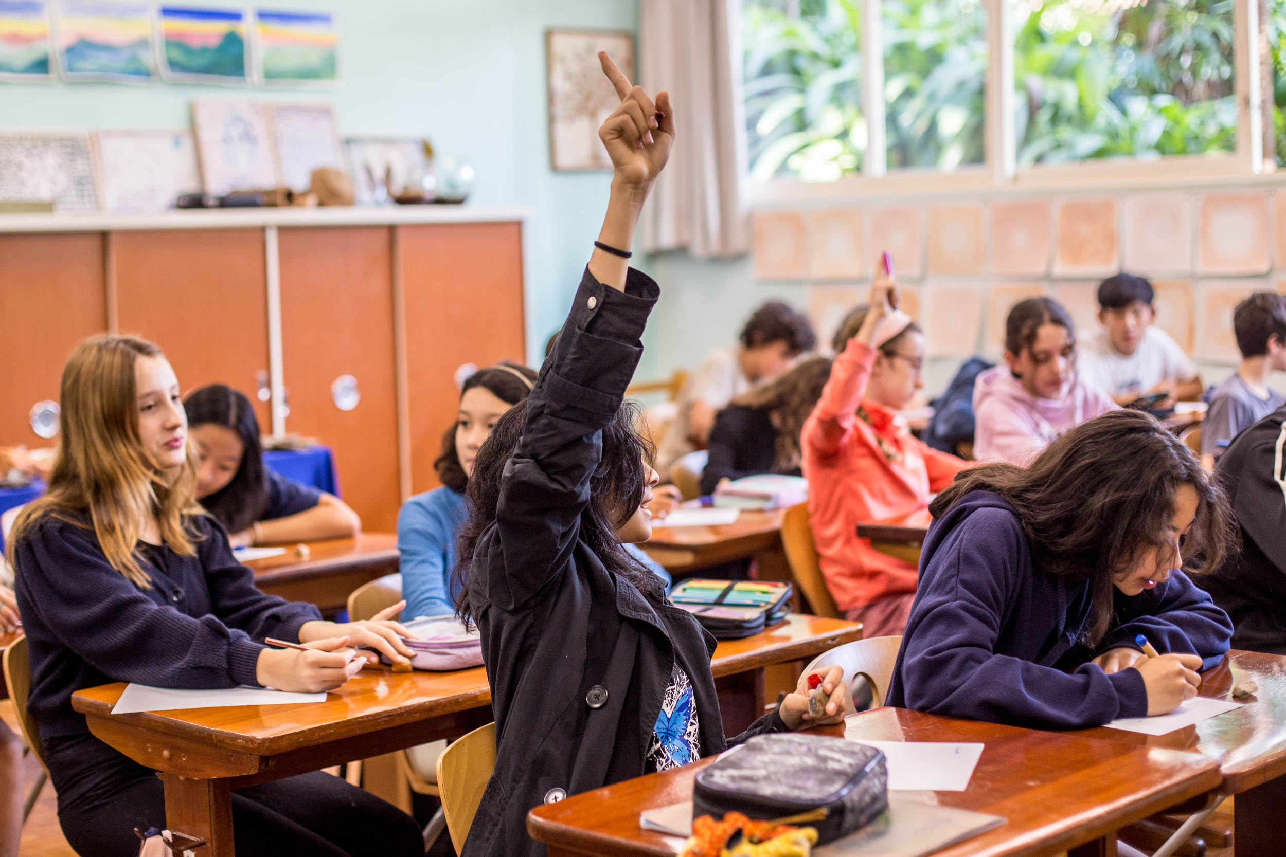 Alunos em sala de aula com uma aluna levantando a mão para participar