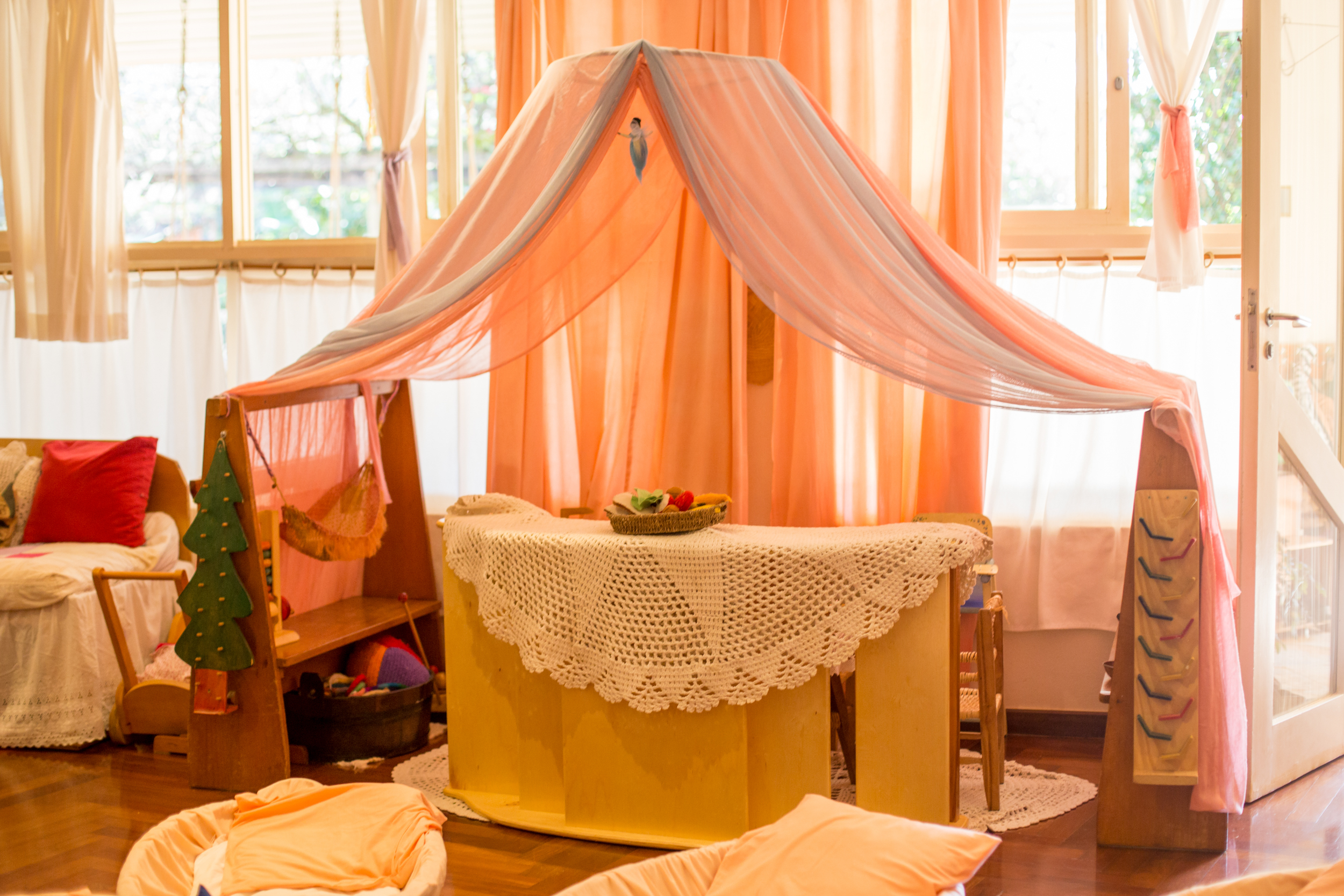 Sala de aula de maternal da Educação Infantil com dossel de tecido sobre mesa, possibilitando brincadeiras imaginativas, janelas grandes e decoração acolhedora.