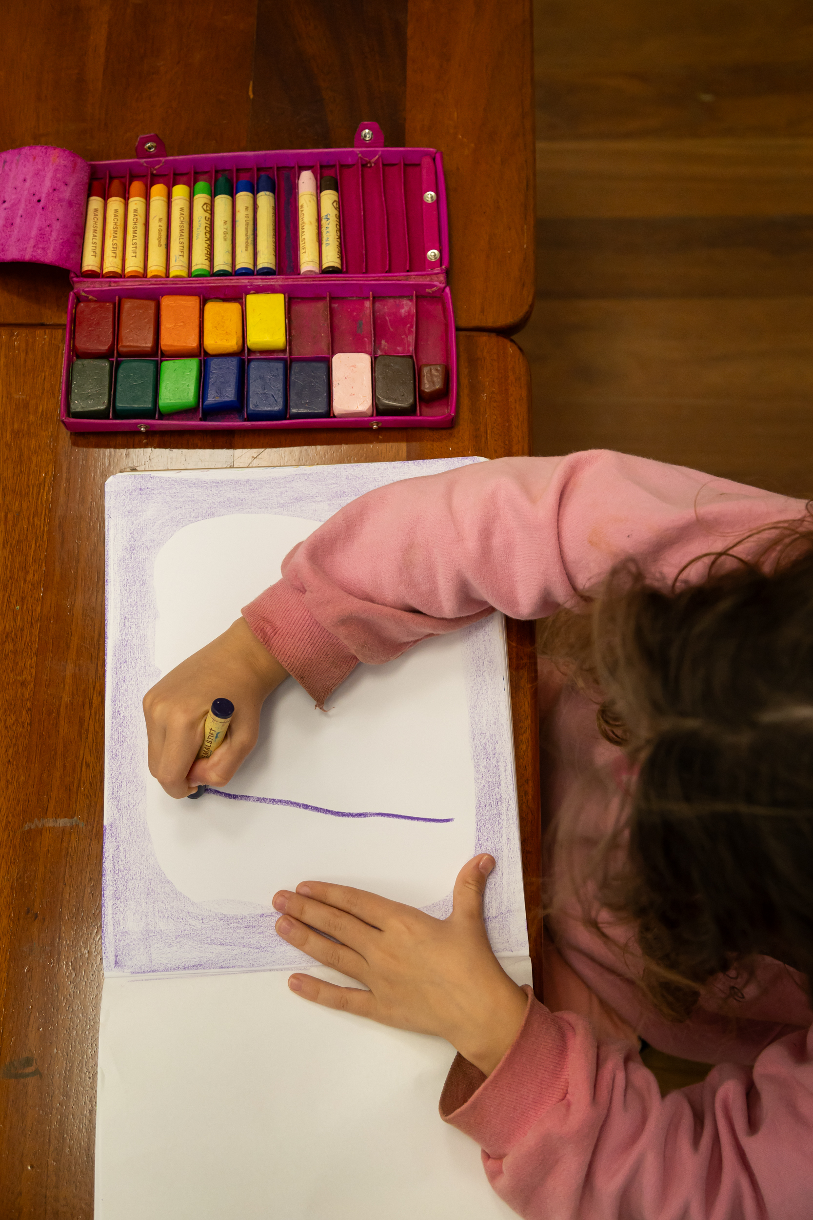 Criança desenhando com giz de cera em caderno e estojo de tintas coloridas