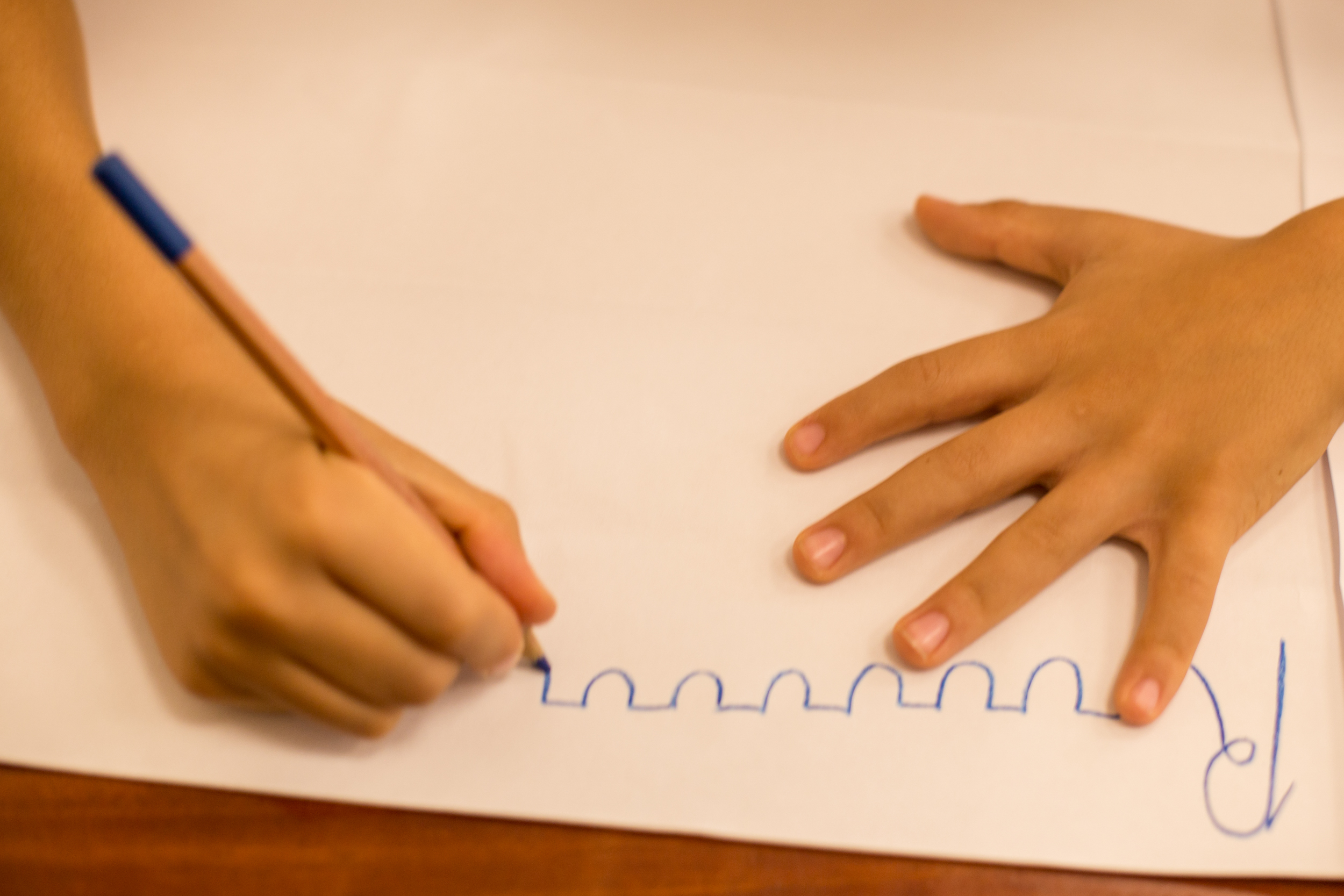 Mãos de criança desenhando formas onduladas com lápis em papel branco em processo de alfabetização em pedagogia Waldorf.