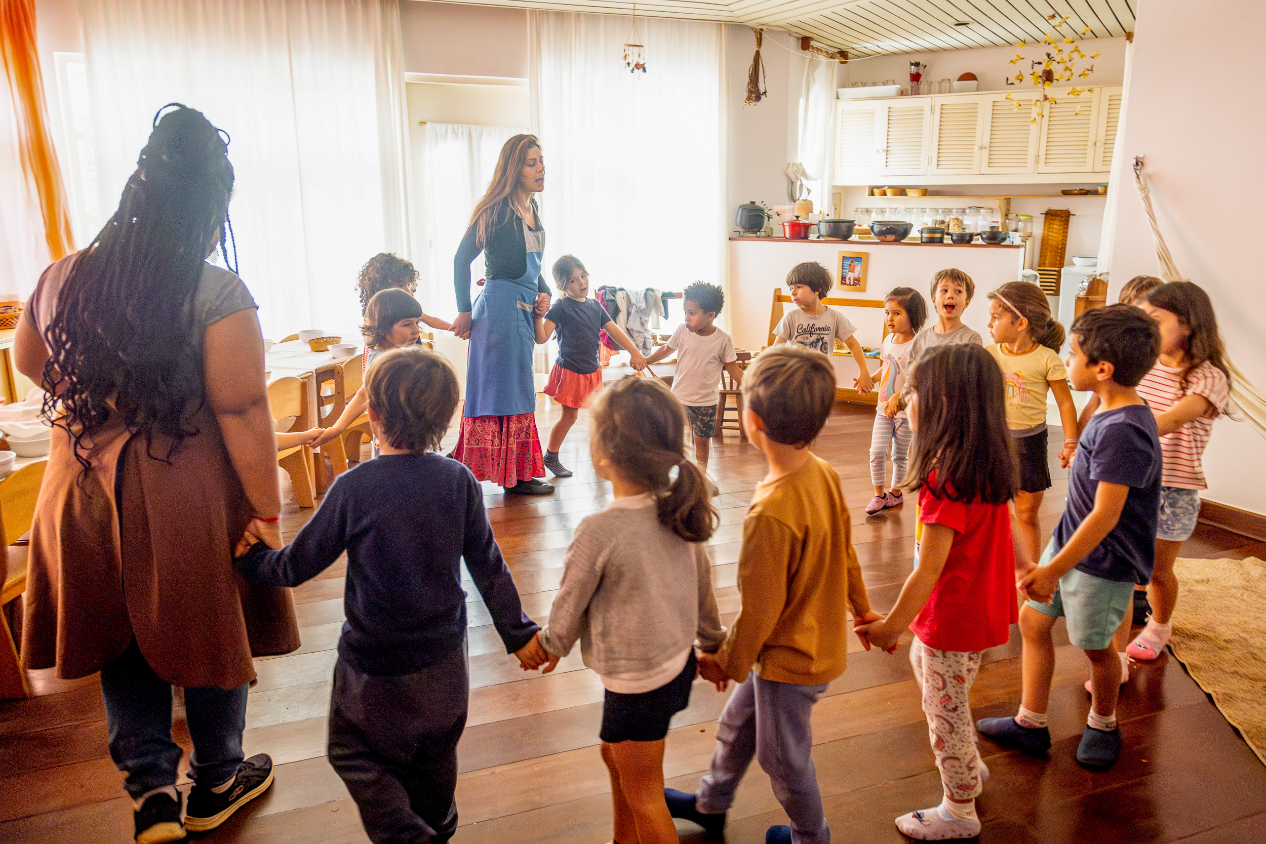Grupo de crianças e professoras de mãos dadas em círculo brincando em sala de aula