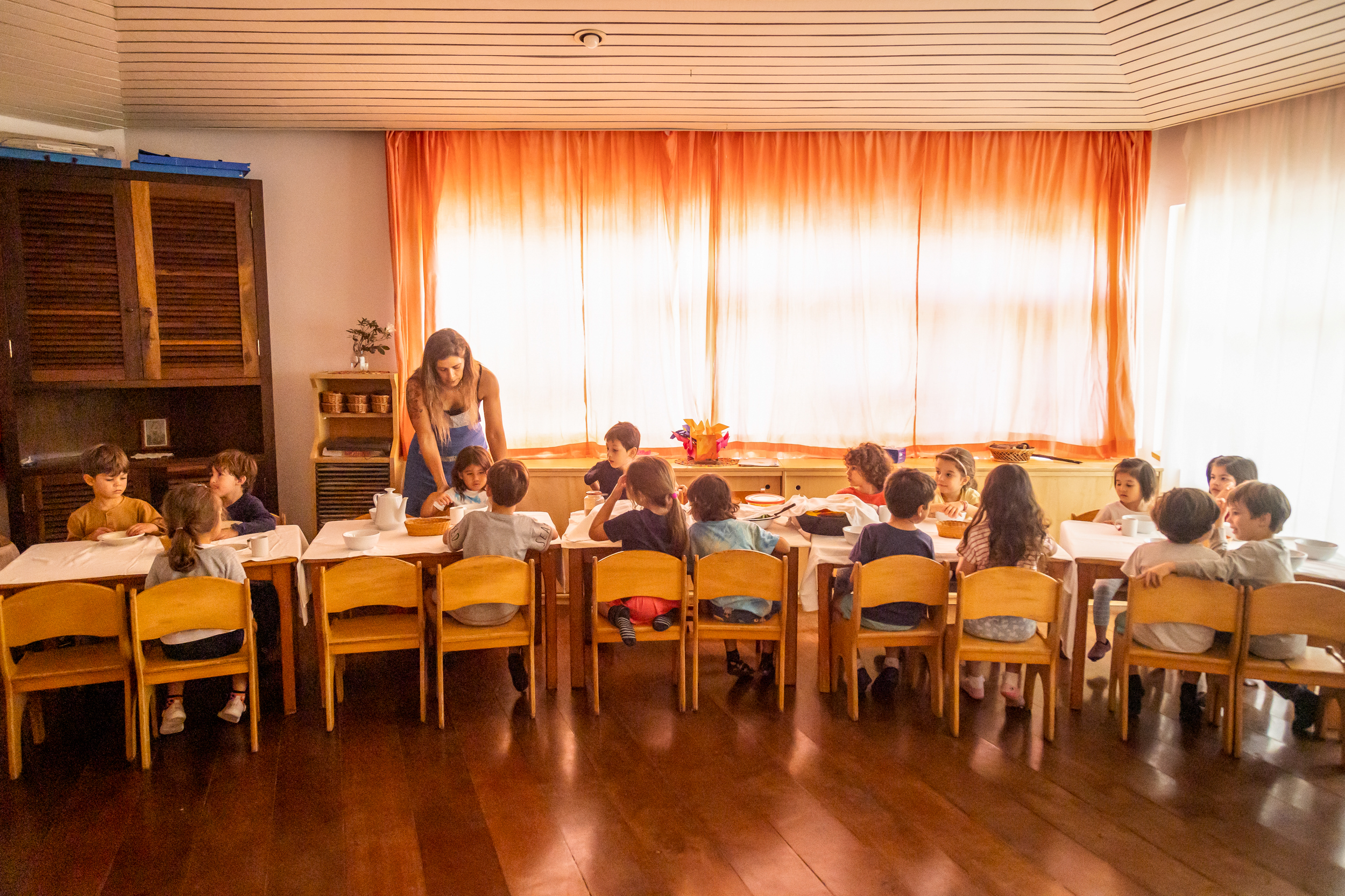 Crianças sentadas em mesas com professora em sala de aula durante refeição