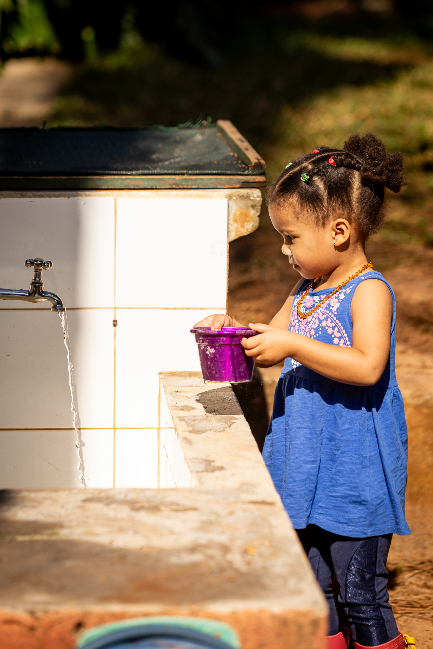 Menina pequena com vestido azul e balde roxo brincando com água de torneira.