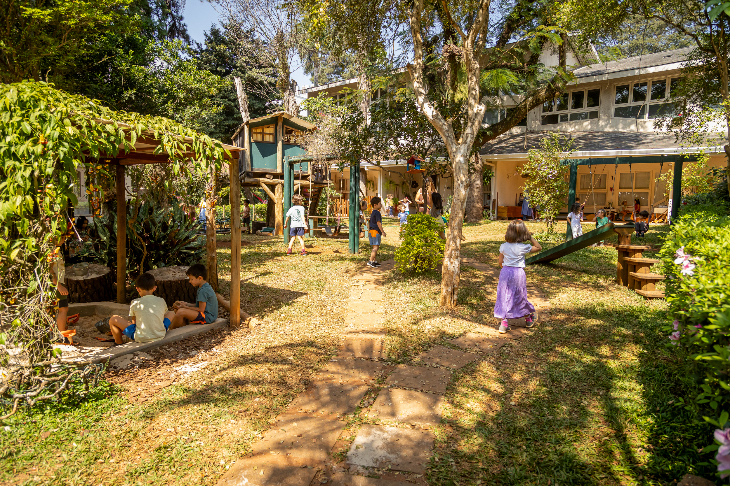 Várias crianças brincando em playground natural com casa na árvore e caixa de areia em escola