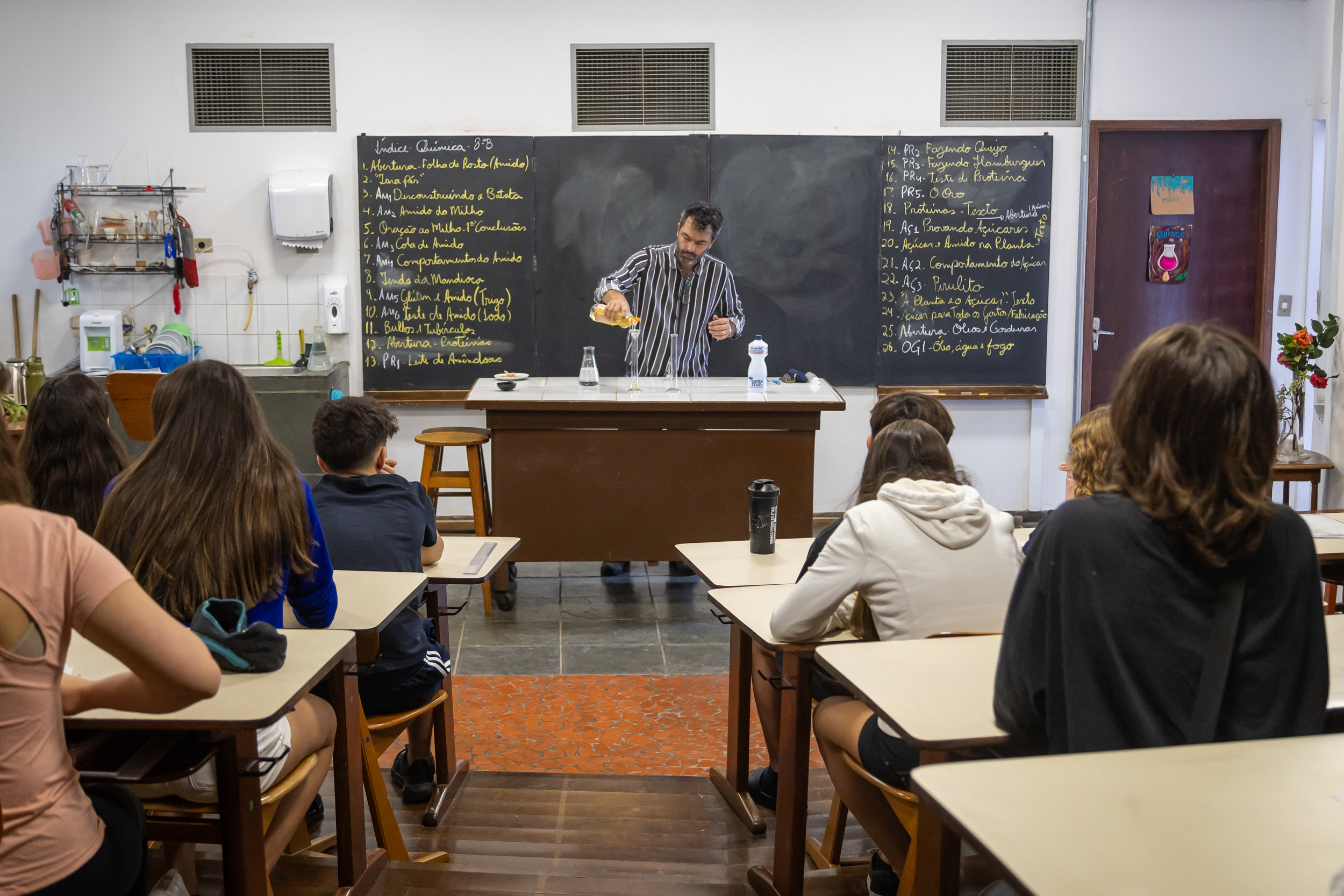 Professor realizando um experimento científico em sala de aula, despejando um líquido em um copo de medição, enquanto os alunos adolescentes o observam com atenção. A imagem ilustra o ensino prático de ciências, como química ou física, incentivando a observação e o interesse em experimentos educacionais.