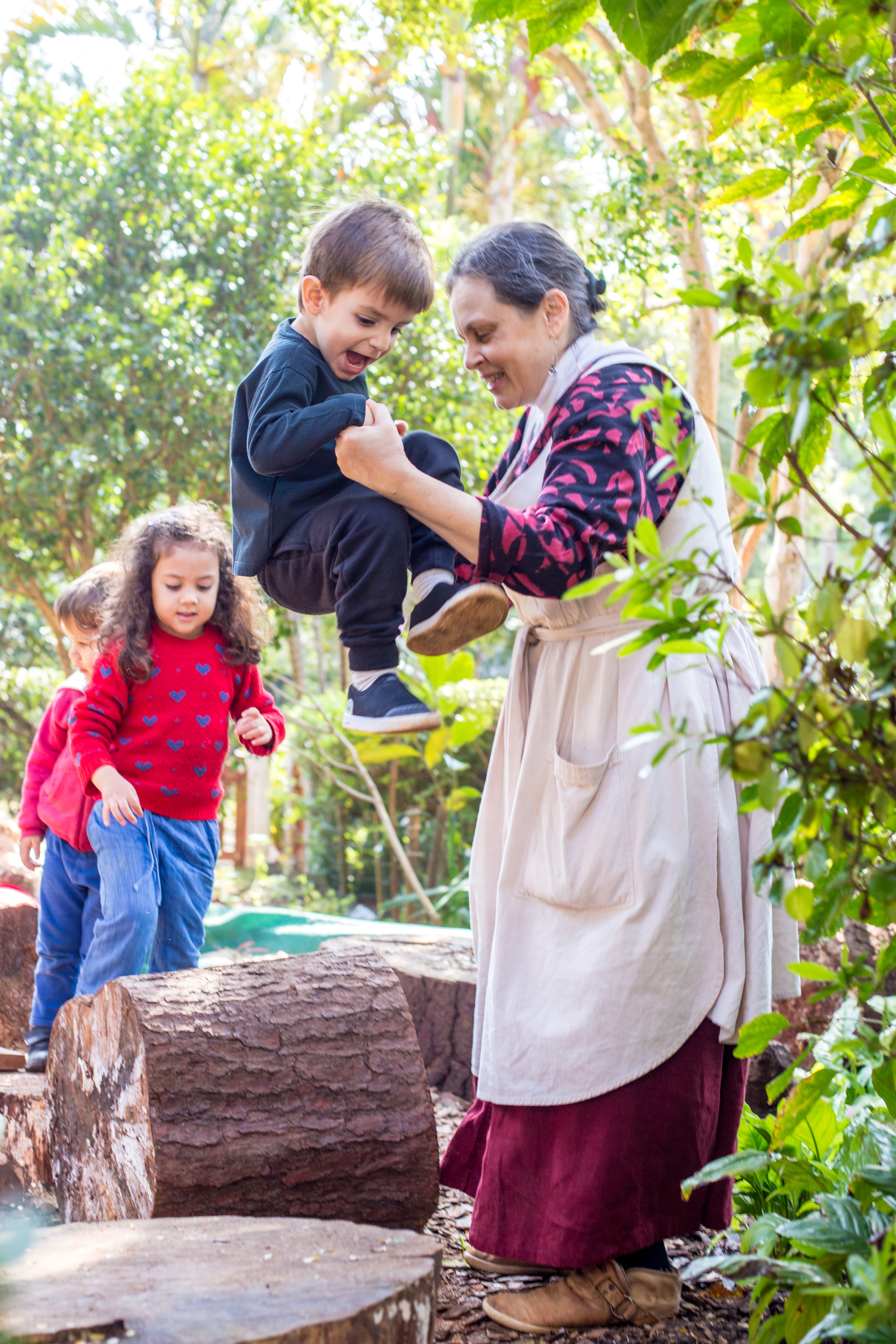 Professora auxiliando menino a pular de tronco de madeira, em playground natural.