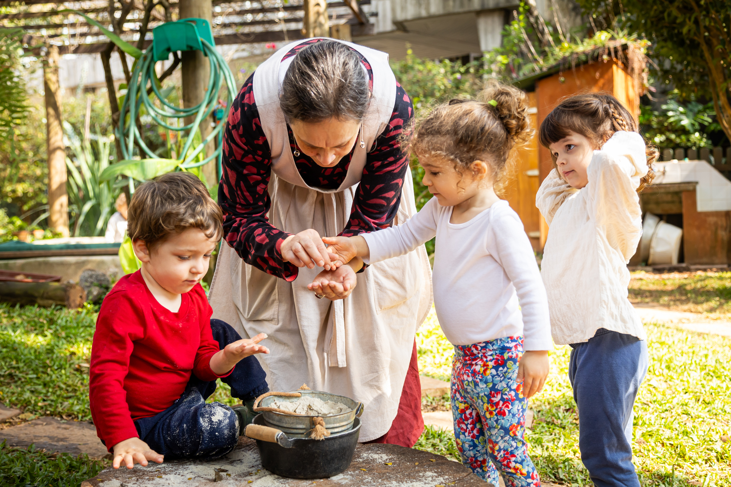 Uma professora de maternal carinhosamente com três crianças pequenas, orientando-as em uma atividade sensorial ao ar livre. As crianças estão reunidas em torno de um pote com terra ou sementes, explorando texturas e desenvolvendo habilidades motoras finas em um ambiente natural e acolhedor de jardim. A imagem ilustra a pedagogia prática e o aprendizado através da experiência na educação infantil.
